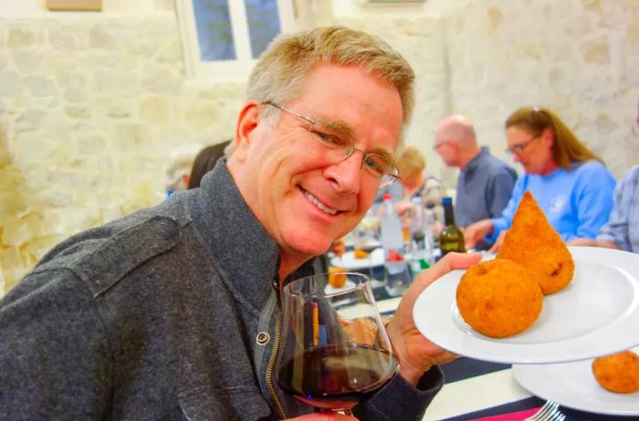 Rick Steves beaming with a plate of crispy snacks and a glass of red wine in Italy