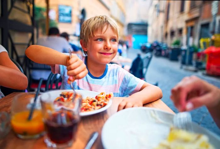 A young boy with blonde hair enjoys pizza with a fork at an outdoor eatery.