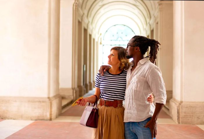 A couple stands within a museum adorned with archways and a beautifully decorated vaulted ceiling in the backdrop.