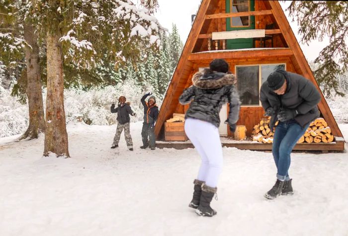 A stylish couple is surprised by two playful children, who launch a flurry of snowballs outside their cozy A-frame cabin.