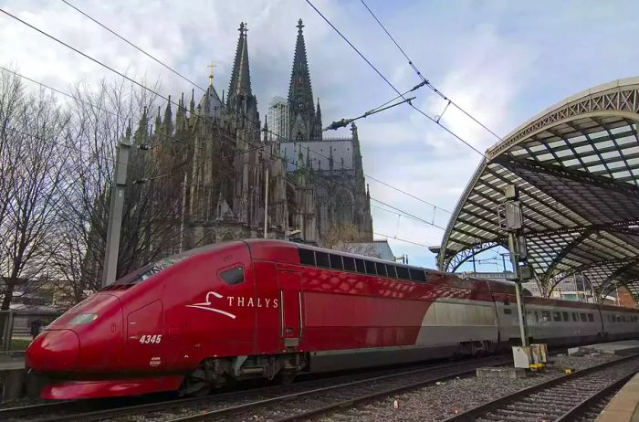 A high-speed Thalys train departs from the central train station in Cologne, Germany, gliding past the iconic Cologne Cathedral, the city's most famous landmark.