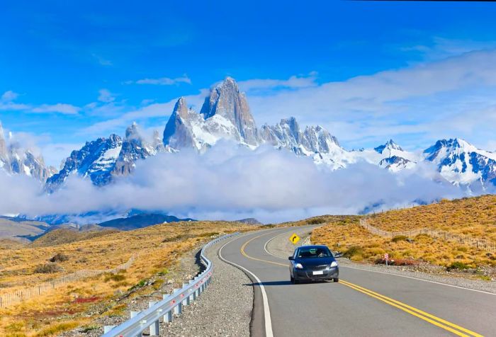 In the background, the majestic Mount Fitz Roy rises, its striking granite silhouette piercing through the mist, crafting a breathtaking scene as a car winds its way along the road away from this natural marvel.