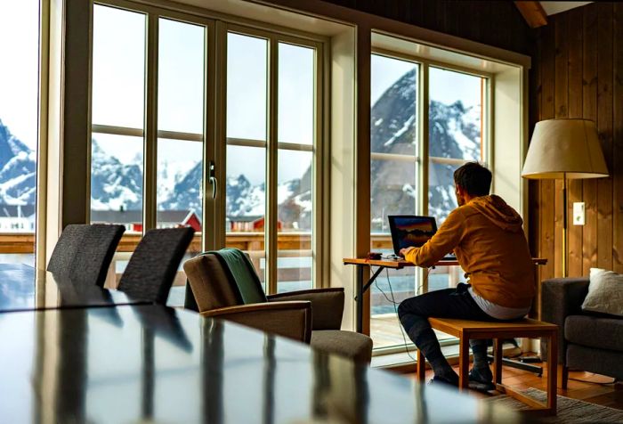 A man in a yellow sweatshirt works on his laptop, enjoying stunning views of the snowy mountains outside.