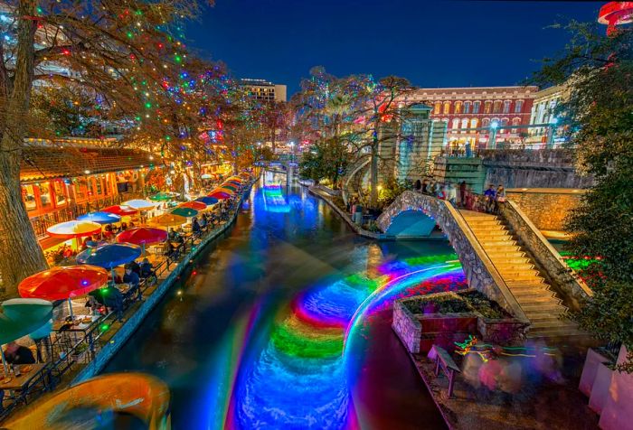 A vibrant promenade with outdoor seating lines a canal adorned with colorful water features.