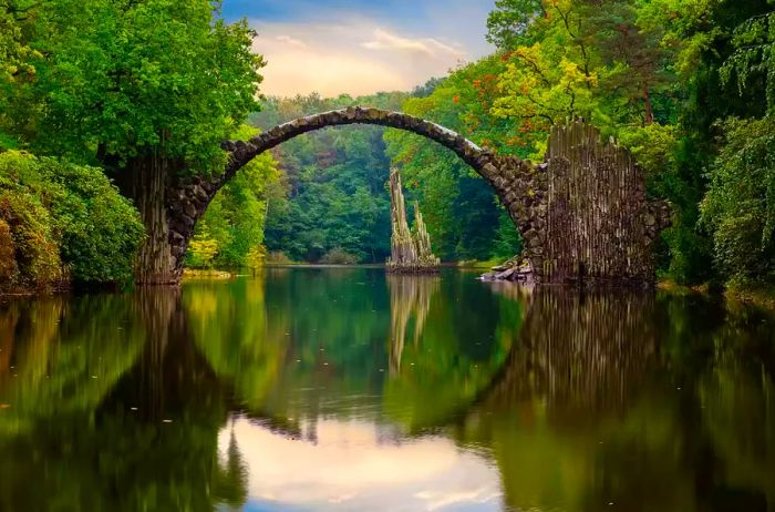 A moody autumn evening casts a clouded sky over the iconic Devil's Bridge in Kromlau, Germany.