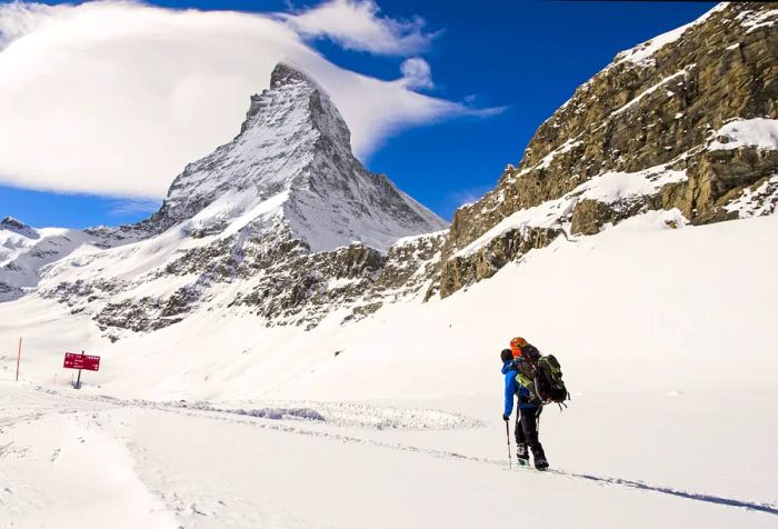 A solitary traveler clad in winter attire carries a bag while trekking along a snowy path beside a majestic mountain range.