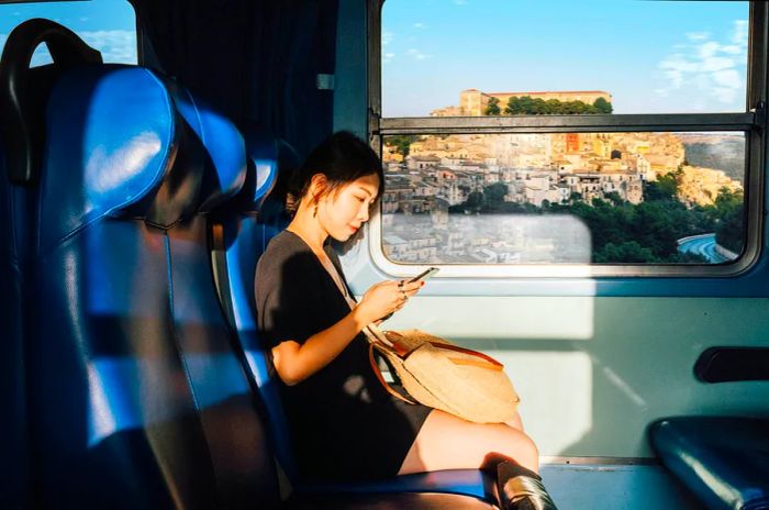 A young East Asian woman is using her phone while on a train in Sicily.