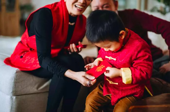 A grandson joyfully receives red envelopes (lai see) from his grandparents while dressed in a traditional red Chinese outfit during the Lunar New Year festivities.