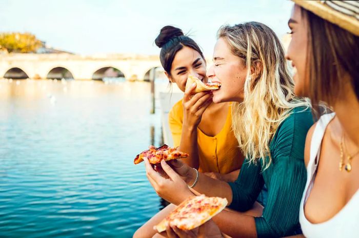 Three friends enjoy pizza topped with cheese and tomato by a riverside in Italy.