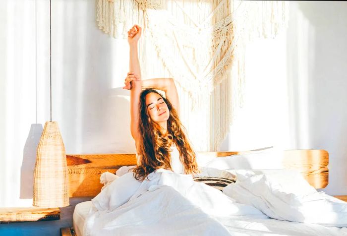 A woman stretches her arms while seated on a bed.