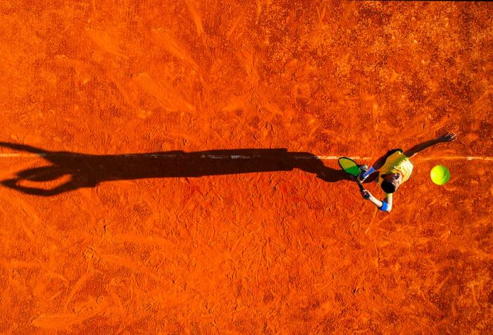 A tennis player serves on a clay court, casting a shadow on the ground.