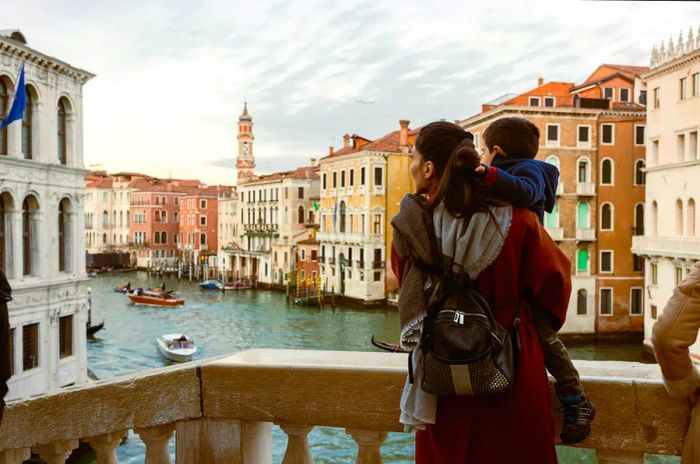 A mother holds her son as they explore Venice. They stand on a bridge overlooking the Grand Canal, with gondolas and boats gliding by. The stunning architecture and breathtaking waterways of Venice create a perfect backdrop for their unforgettable adventure.