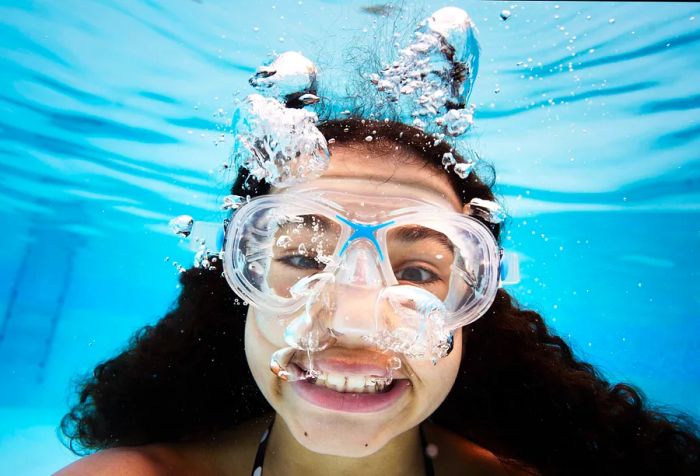 A teen girl smiles at the camera while blowing bubbles underwater.