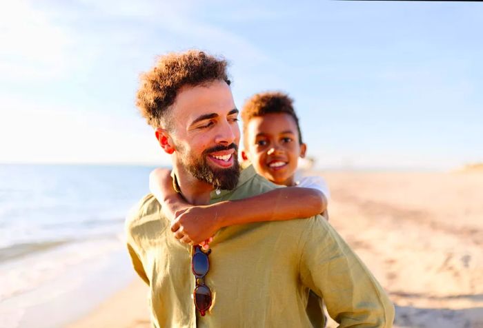 A man walks along the beach carrying a little boy on his back.