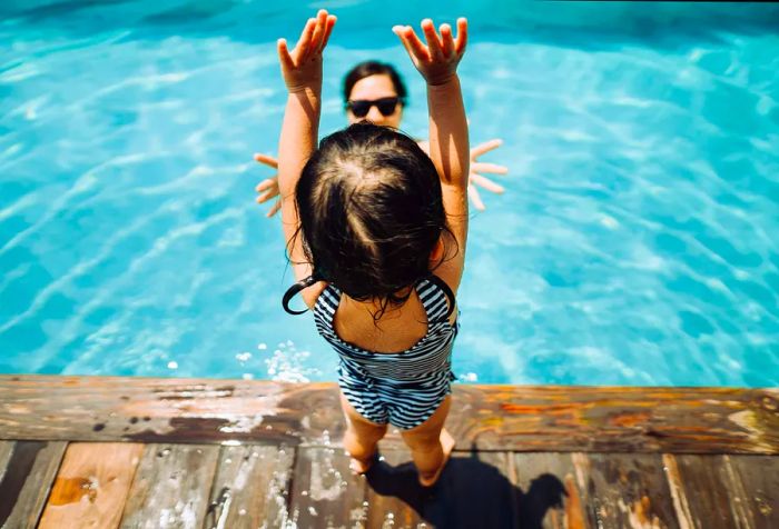 An excited toddler girl stands by the pool with her arms raised, watching her mother swim.