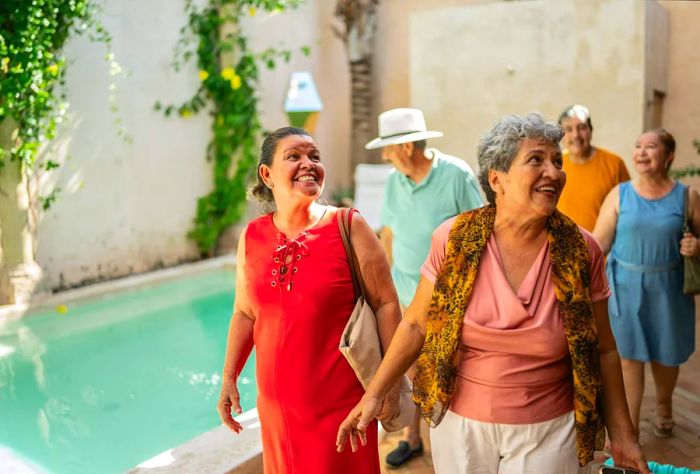 A group of older friends walks past a sparkling pool as they arrive at their hotel.