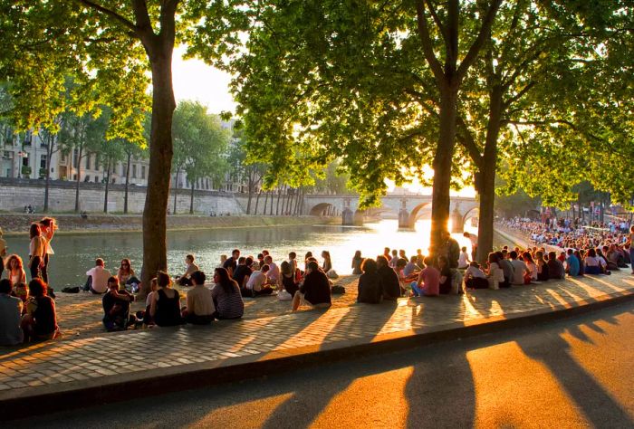 Individuals relaxing under the shade of trees beside a riverbank.