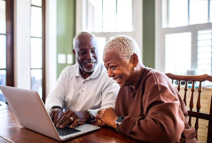 An elderly couple happily using a laptop on a wooden table.