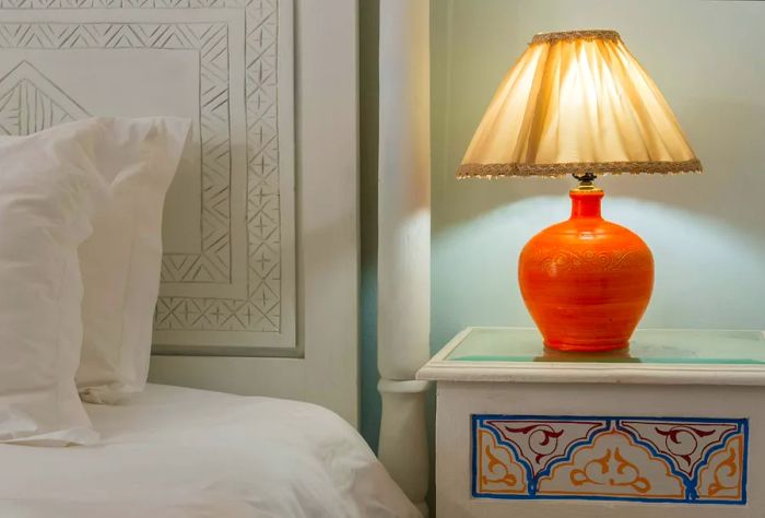 A glowing lampshade perched on a small table next to a white bed.