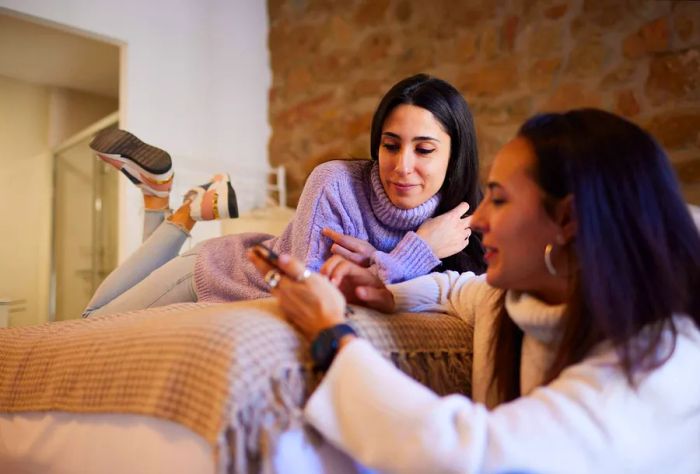 One friend lies on the bed facing down while the other sits on the floor, sharing a smartphone screen, both fully absorbed in what they’re viewing together.