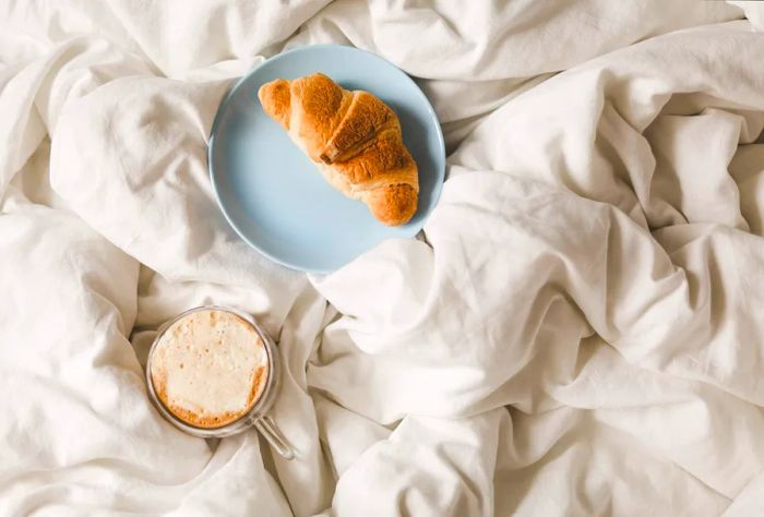 A croissant elegantly placed on a blue plate next to a cup of coffee, all resting on a white bed.