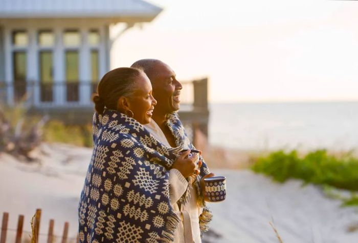 An elderly couple wrapped in a knitted shawl smiles as they enjoy their time on the beach.