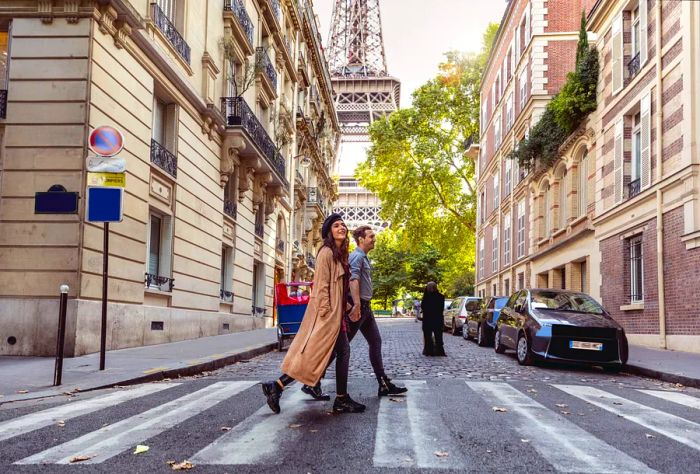 A casually dressed couple strolls across a crosswalk with the Eiffel Tower looming in the background.