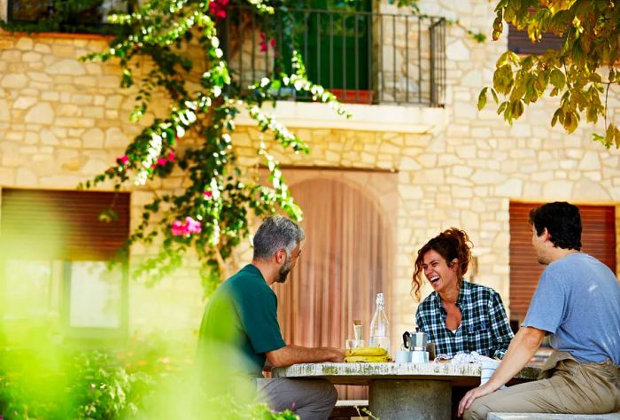 A cheerful group of coworkers enjoys a meal together outdoors around a round table, with a picturesque stone house in the background.