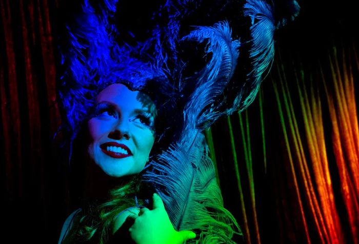 A showgirl in costume and stage makeup flashes a sultry smile while preparing backstage for her performance. She wears a dazzling blue sequin outfit topped with a feather headdress, illuminated by vibrant lighting.