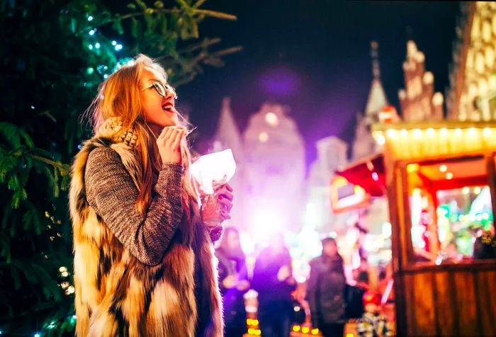 A blonde woman in fur and glasses beams as she gazes at the holiday lights while enjoying a snack.