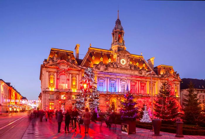 A stunning Renaissance-style building glowing in various colors, adorned with numerous illuminated Christmas trees in its garden.