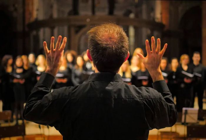 A choirmaster gestures passionately against a bokeh background of a crowd.