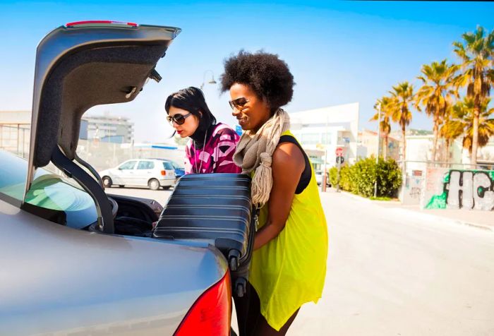 Two women loading heavy luggage into an open car trunk in a parking lot.