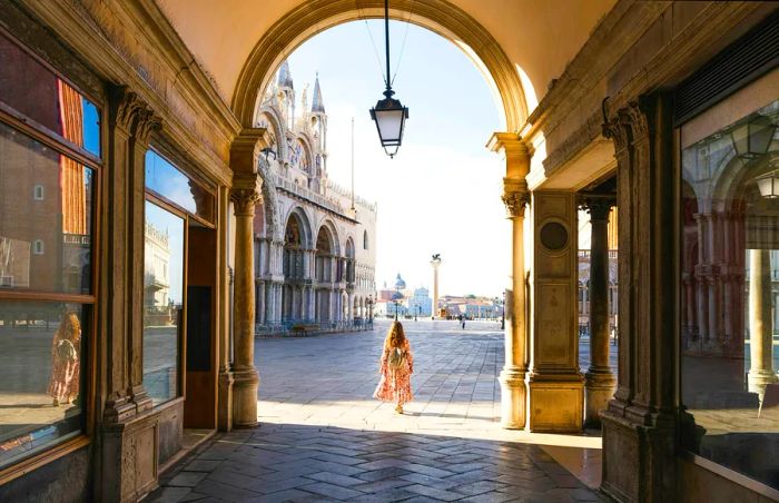 A woman strolls through the arcade leading to St Mark's Square, with St Mark's Cathedral in the backdrop. Venice, Veneto region, Italy.