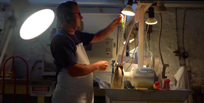 A craftsman in a workshop lifts a piece of greenstone toward the light, preparing to carve it.