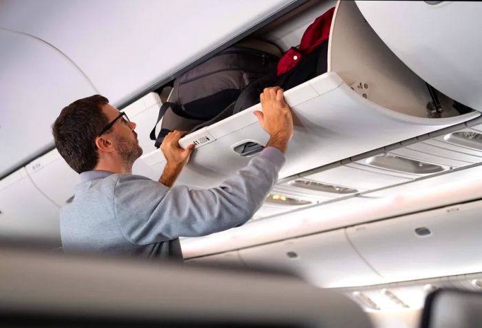 A man securing his luggage in the overhead bin of the airplane.