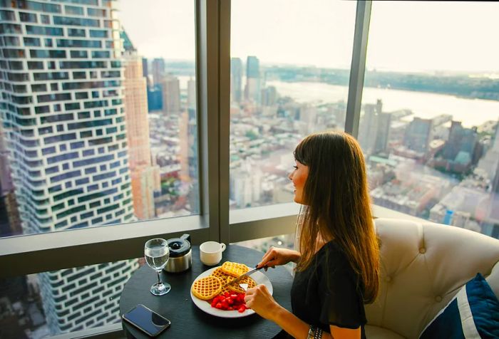 A woman enjoying waffles and strawberries at a table, gazing out at the city through large glass windows.