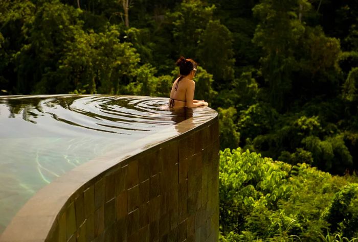 A person in swimwear relaxes by the pool, gazing at the jungle beyond.