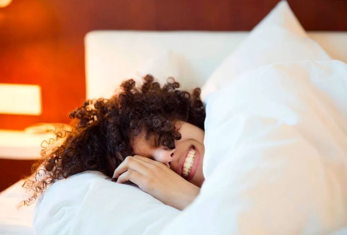 A woman with curly hair beams while cozying up under a blanket.