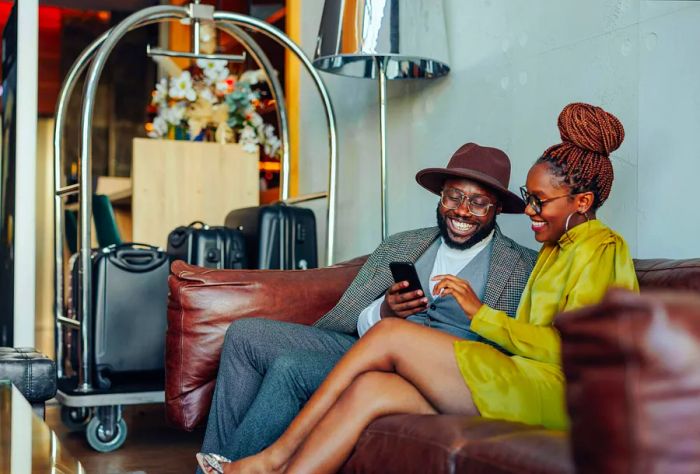 A couple is relaxing in the hotel, waiting to be escorted to their luxurious suite. They sit comfortably on a sofa, luggage on a cart beside them, while they browse on a smartphone.