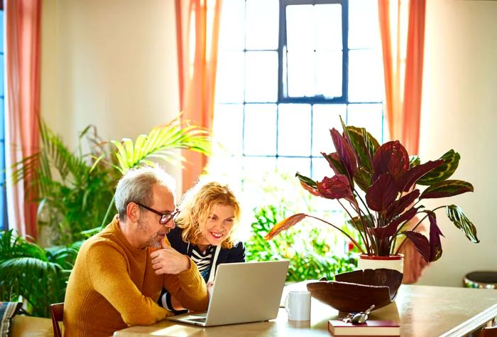 An older couple sits side by side, absorbed in their computer, surrounded by lush, colorful plants.