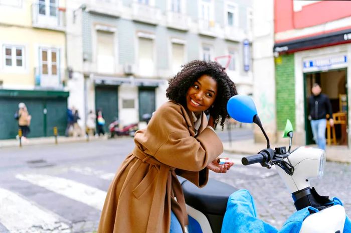 A woman on a scooter in Lisbon, Portugal, adjusting her hair while looking in a mirror 1478410737 20-30 years, adults, of African descent, dark hair, female, female African American, grande lisboa, happy, leisure, millennials, POC, three-quarter length, young adult