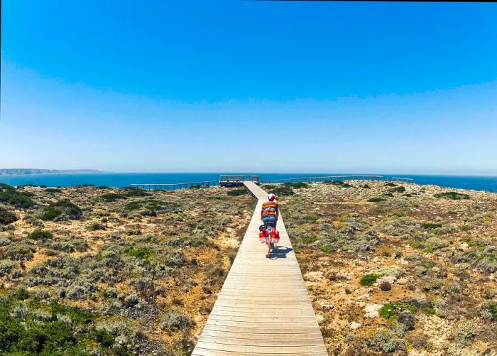 A young woman pedals a bicycle along a narrow wooden boardwalk in Carrapateira, Portugal, with the blue sea visible in the distance.