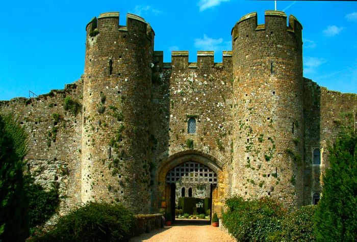 An entrance to an ancient castle featuring a portcullis and flanked by two towers, all surrounded by lush greenery.