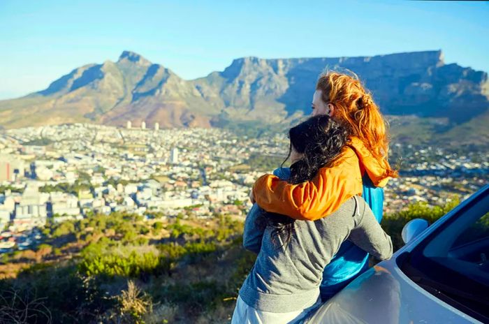 Two women embrace beside their car with Table Mountain towering in the background