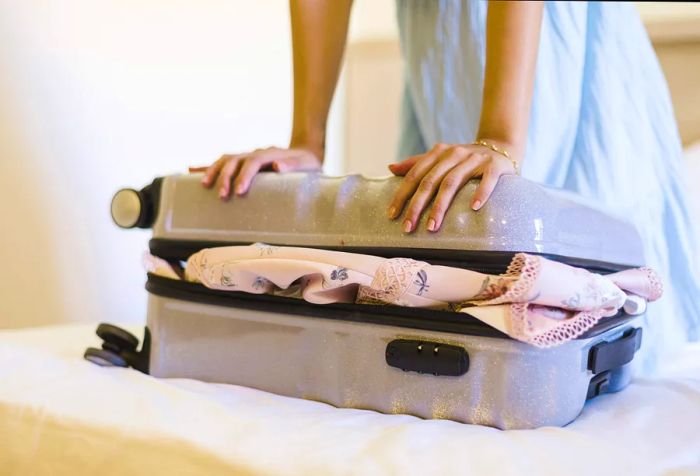 Hands attempting to close a suitcase filled with clothes.