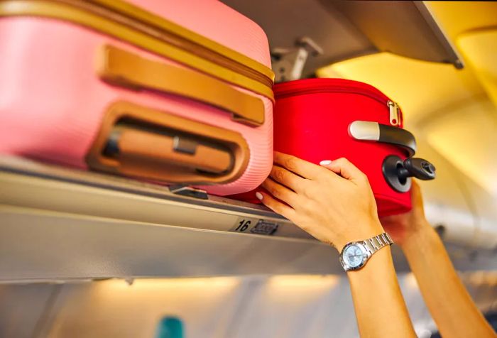 A woman is trying to fit a red suitcase into the overhead compartment's tight space.