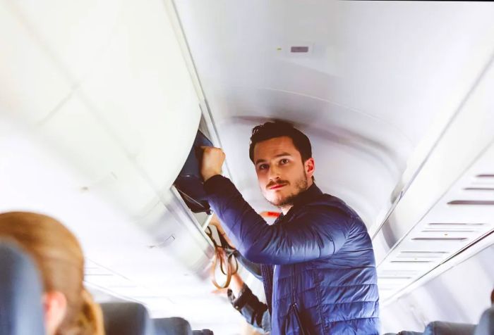 A tall, stylish man placing his luggage into the overhead compartment.