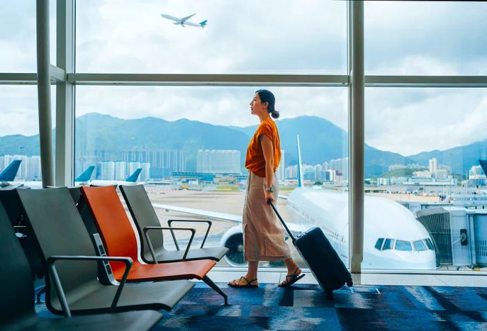 An Asian woman walking through an airport terminal with a suitcase, passing by a window.