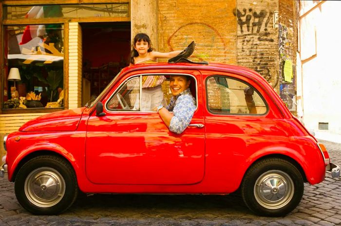 A cheerful father and daughter enjoying a ride in a Fiat 500 in Italy, with the daughter playfully standing outside the sunroof.